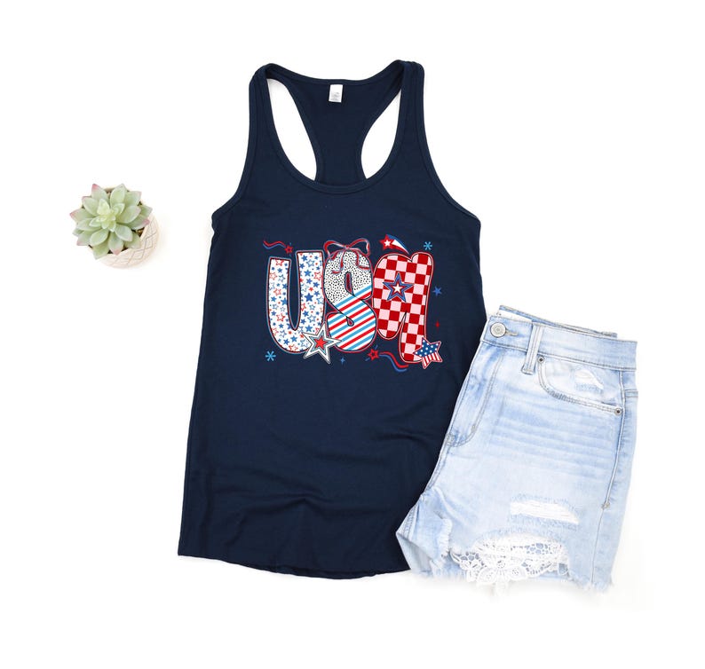 May include: A navy blue tank top with the word "USA" in a patriotic design. The letters feature patterns of stars, stripes, and checkered elements in red, white, and blue. Light blue denim shorts are next to the tank top.