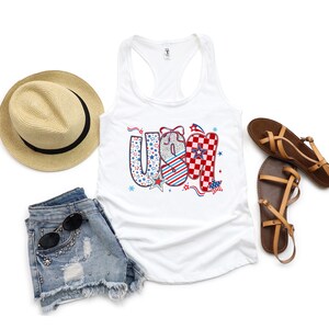 May include: A white tank top with the word "USA" in a patriotic design, paired with denim shorts, a straw hat, and brown sandals. The "USA" lettering features stars, stripes, and a checkered pattern. Sunglasses are on the shorts.
