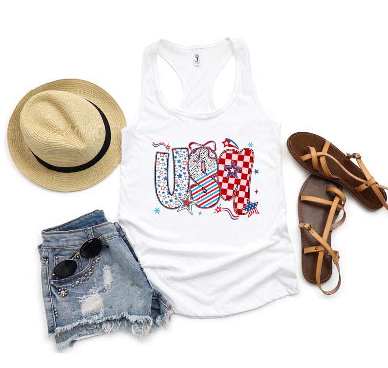 May include: A white tank top with the word "USA" in a patriotic design, paired with denim shorts, a straw hat, and brown sandals. The "USA" lettering features stars, stripes, and a checkered pattern. Sunglasses are on the shorts.