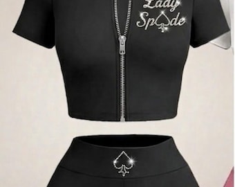 May include: A black two-piece set consisting of a short-sleeved zip-up crop top and high-waisted shorts. The top features the text Lady Spade in rhinestones. A matching black headband with a rhinestone spade symbol is included.