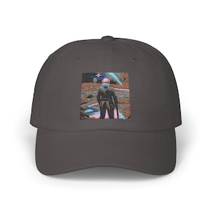 May include: A charcoal gray baseball cap featuring an embroidered image of a figure in a landscape. The artwork depicts a person in a dark outfit with a weapon, standing in a desert-like environment. The cap has a curved brim and a structured crown.