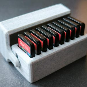 May include: A white plastic case with a hinged lid, designed to hold and store Nintendo Switch game cartridges. The case is filled with 12 game cartridges, each with a black and red design.