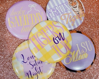 May include: Five circular badges with different designs and text. The badges feature phrases such as "Lucky Gameday But" and "LSU Mom." One badge has a tiger illustration. Colours include purple, yellow, and white.
