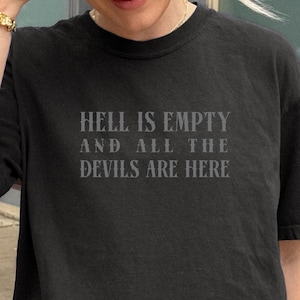 Puede incluir: Camiseta negra con el texto "HELL IS EMPTY AND ALL THE DEVILS ARE HERE" en gris. La camiseta tiene cuello redondo y mangas cortas. La camiseta está siendo usada.