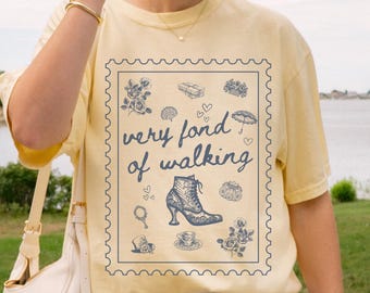 May include: A light yellow t-shirt with a graphic that reads "very fond of walking" in a stamp-like design. The graphic includes illustrations of flowers, a boot, and other vintage-style elements. The shirt is paired with white shorts.