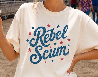 May include: A cream-colored short-sleeve t-shirt featuring the text Rebel Scum in a blue script font, surrounded by small red and blue stars. The shirt is paired with blue denim jeans, creating a casual look with a patriotic color palette.