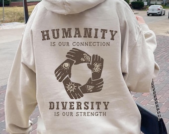 May include: A beige hoodie with the text "HUMANITY IS OUR CONNECTION" and "DIVERSITY IS OUR STRENGTH". The back of the hoodie features a graphic of four hands forming a circle. The hands have sun and eye designs.