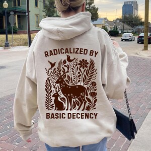 May include: Beige hoodie with a brown graphic featuring a deer, two smaller animals, and floral elements. The text "RADICALIZED BY BASIC DECENCY" is printed above and below the graphic. The hoodie has a hood and long sleeves.