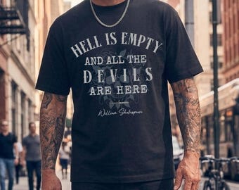 May include: Black t-shirt with the quote "Hell is Empty and All the Devils Are Here" in white lettering. The design includes a skull and floral elements, with "William Shakespeare" below. The shirt is worn by a person in an urban setting.