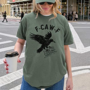 May include: Olive green t-shirt with a black raven graphic and the text "F-CAW-F". The raven is perched on a stack of books. The shirt has short sleeves and a crew neck.