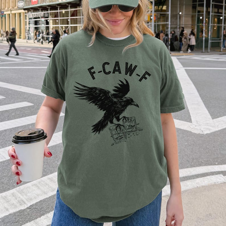 May include: Olive green t-shirt with a black raven graphic and the text "F-CAW-F". The raven is perched on a stack of books. The shirt has short sleeves and a crew neck.