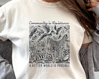 May include: A white t-shirt with a black and white illustration of animals and plants. The text "Community is Resistance" and "A Better World is Possible" is printed on the shirt.
