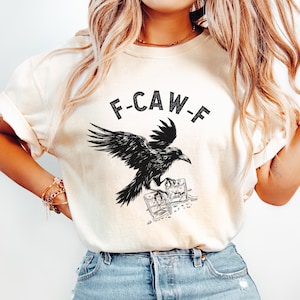 May include: Cream-colored t-shirt featuring a black raven illustration with the text "F-CAW-F". The raven is depicted with outstretched wings, perched above two small square glasses. The shirt is made of a soft material.