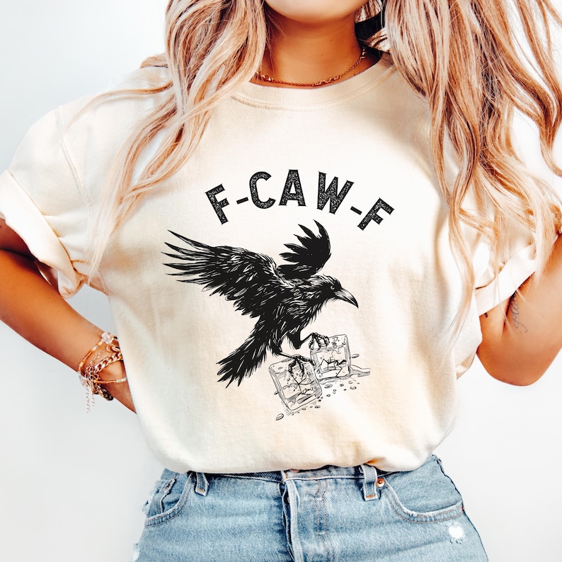May include: Cream-colored t-shirt featuring a black raven illustration with the text "F-CAW-F". The raven is depicted with outstretched wings, perched above two small square glasses. The shirt is made of a soft material.