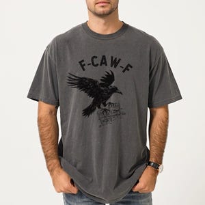 May include: Dark gray t-shirt with a black raven graphic and the text "F-CAW-F". The raven is perched on a stack of books. The shirt has short sleeves and a crew neck.