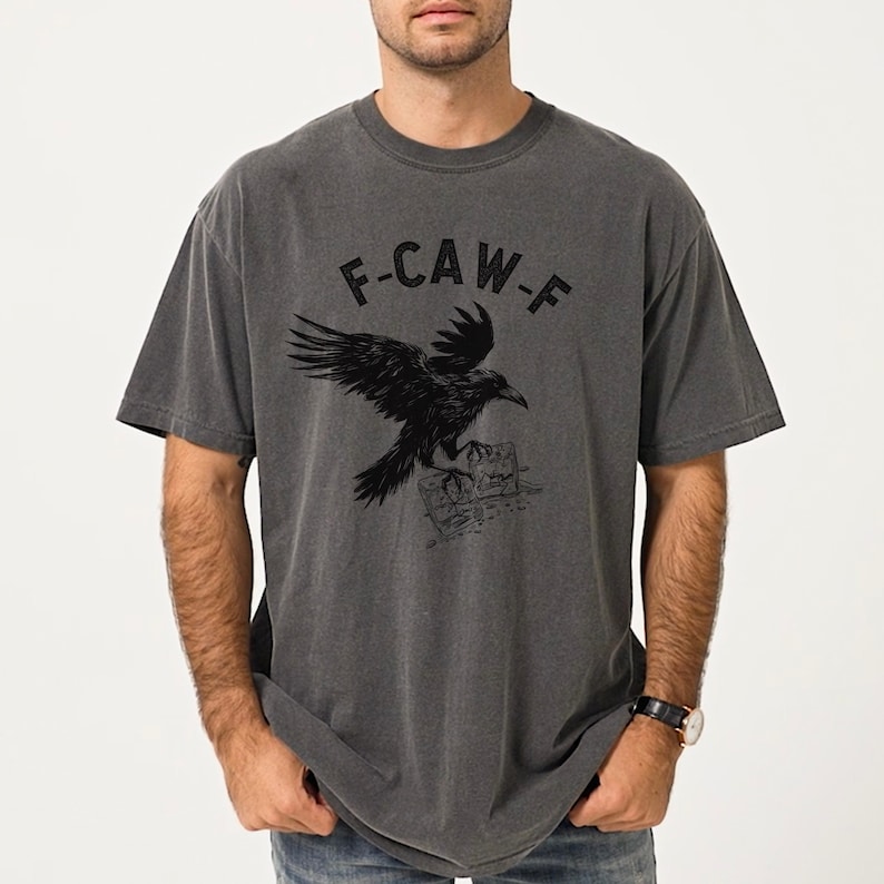 May include: Dark gray t-shirt with a black raven graphic and the text "F-CAW-F". The raven is perched on a stack of books. The shirt has short sleeves and a crew neck.