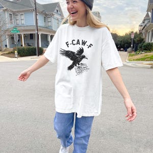 May include: White t-shirt with a black raven graphic and the text "F-CAW-F". The shirt is paired with blue jeans. The raven is perched on a detailed illustration. The person is wearing a gray beanie.