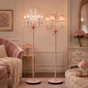 May include: Two pink floor lamps with crystal chandeliers and pink lampshades light up a room. The lamps have a slender, pink metal pole and a round base. The room features a pink sofa, a gold-framed mirror, and a decorative table with a bouquet of pink roses.