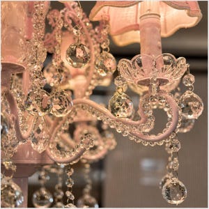 May include: Close-up of a pink chandelier with crystal accents. The chandelier features multiple tiers of sparkling crystal pendants and teardrops, with a pink painted metal frame and a pink lampshade.