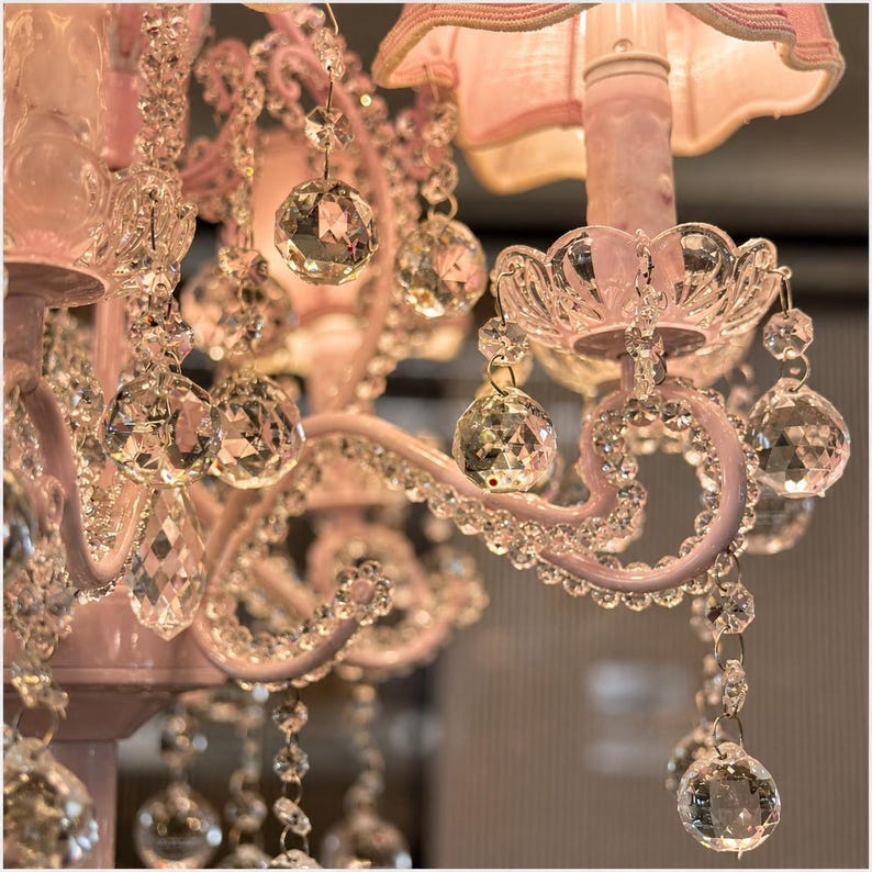 May include: Close-up of a pink chandelier with crystal accents. The chandelier features multiple tiers of sparkling crystal pendants and teardrops, with a pink painted metal frame and a pink lampshade.