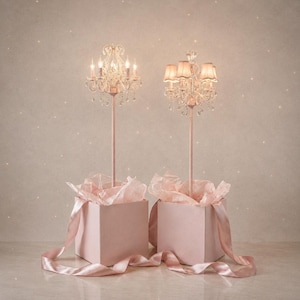 May include: Two pink decorative chandeliers with crystal detailing and lit candles. Each chandelier is mounted on a pink stand, presented in a pink gift box with pink tissue paper and a satin ribbon. The background is a soft, speckled pink.