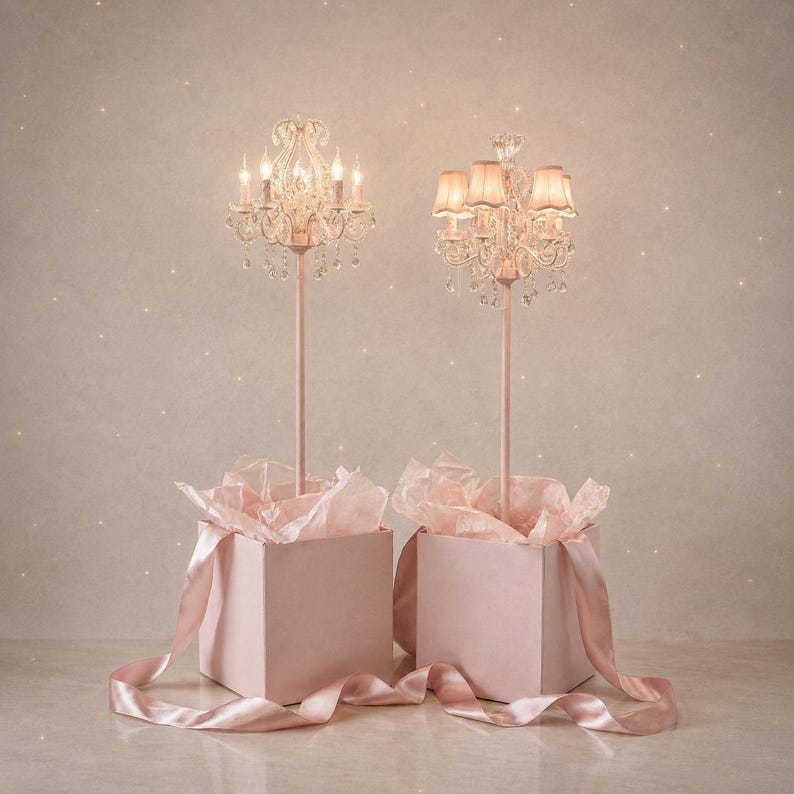 May include: Two pink decorative chandeliers with crystal detailing and lit candles. Each chandelier is mounted on a pink stand, presented in a pink gift box with pink tissue paper and a satin ribbon. The background is a soft, speckled pink.