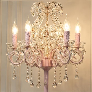 May include: A pink and crystal chandelier with five candle-shaped lights. The chandelier features a decorative design with crystal accents and hanging crystal droplets. The light fixtures are pink, and the overall design is ornate and elegant.