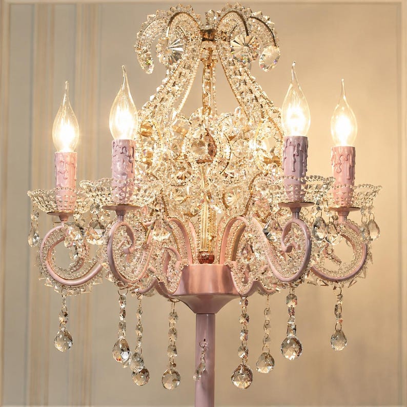 May include: A pink and crystal chandelier with five candle-shaped lights. The chandelier features a decorative design with crystal accents and hanging crystal droplets. The light fixtures are pink, and the overall design is ornate and elegant.