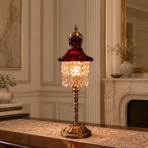 May include: A table lamp featuring a deep red glass shade with a fluted design, accented by a cascading fringe of clear crystal-like beads and teardrop prisms. The lamp has a brass-toned metal base and a slender, decorative column with a pull chain.