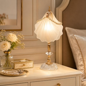 May include: A table lamp featuring a frosted glass shade shaped like a seashell, adorned with a string of white pearls. The lamp has a gold-toned metal stem and a clear, faceted crystal base that reflects light, creating a warm, soft glow.