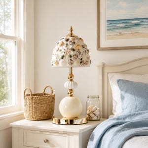 May include: A decorative table lamp with a seashell-covered shade, a cream-colored base, and gold accents. The lamp sits on a white nightstand next to a woven basket and a jar filled with seashells. A framed beach scene hangs on the wall.