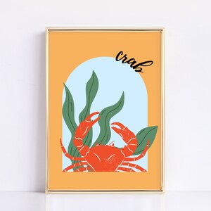 Op de afbeelding: Een ingelijst print met een gele achtergrond en een blauwe boog. De boog heeft een rode krab met groen zeewier. De tekst "crabs" is in zwarte cursieve letters boven de boog geschreven.