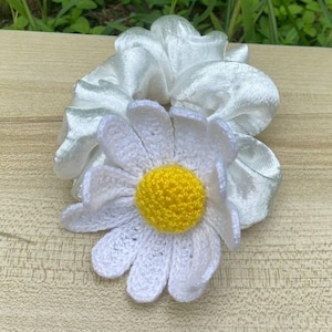 以下が含まれることがあります： 白いサテンのシュシュと、かぎ針編みのデイジーの花。 デイジーは白い花びらと黄色の中心を持っています。 シュシュと花は、明るい茶色の木製の表面にあります。