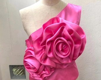 Peut inclure: Un haut asymétrique rose vif orné de deux grandes roses en tissu en relief sur le devant. Le vêtement est confectionné dans une matière satinée lisse et brillante, présentant un corsage ajusté et une fermeture éclair latérale.