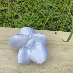 May include: A white satin flower with five petals and a rounded center. The flower is placed on a light brown wooden surface, with green grass and foliage visible in the background. The petals have a soft, textured appearance. Handmade Fluffy Floral Scrunchie: Long Hair Tie