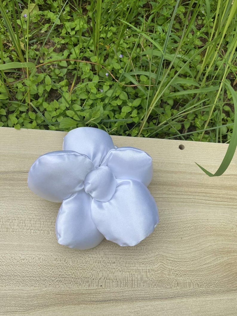 May include: A white satin flower with five petals and a rounded center. The flower is placed on a light brown wooden surface, with green grass and foliage visible in the background. The petals have a soft, textured appearance. Handmade Fluffy Floral Scrunchie: Long Hair Tie