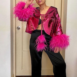 May include: A magenta sequined jacket with bright pink feather trim on the cuffs. The jacket is open in the front and tied with a magenta ribbon. The model is wearing black satin trousers.