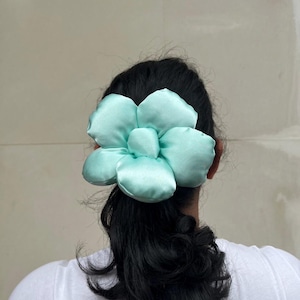 A large, light green flower-shaped hair scrunchie. The scrunchie is made of a soft, satin-like material and is attached to the back of a person's head, secured in their dark hair. The hair is styled in a low ponytail, with the accessory positioned above the elastic. Handmade Fluffy Floral Scrunchie: Long Hair Tie