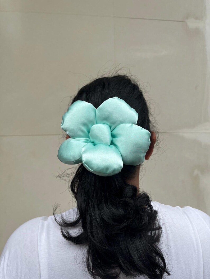 A large, light green flower-shaped hair scrunchie. The scrunchie is made of a soft, satin-like material and is attached to the back of a person's head, secured in their dark hair. The hair is styled in a low ponytail, with the accessory positioned above the elastic. Handmade Fluffy Floral Scrunchie: Long Hair Tie