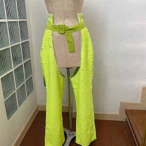 Puede incluir: Chaps de color verde neón con cinturón y hebilla a juego, expuestos en un maniquí. Los chaps tienen un aspecto texturizado, posiblemente con lentejuelas. El cinturón es de color verde neón liso.