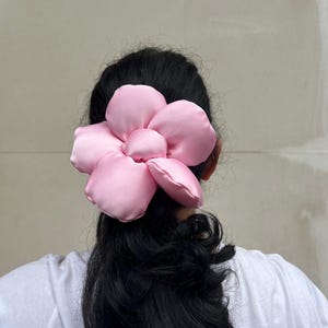 May include: A large, pink flower scrunchie. The hair scrunchie is made of a soft, satin-like material. The flower has multiple petals and a rounded center. The scrunchies is worn on the back of a person's head, with black hair visible. Handmade Fluffy Floral Scrunchie: Long Hair Tie