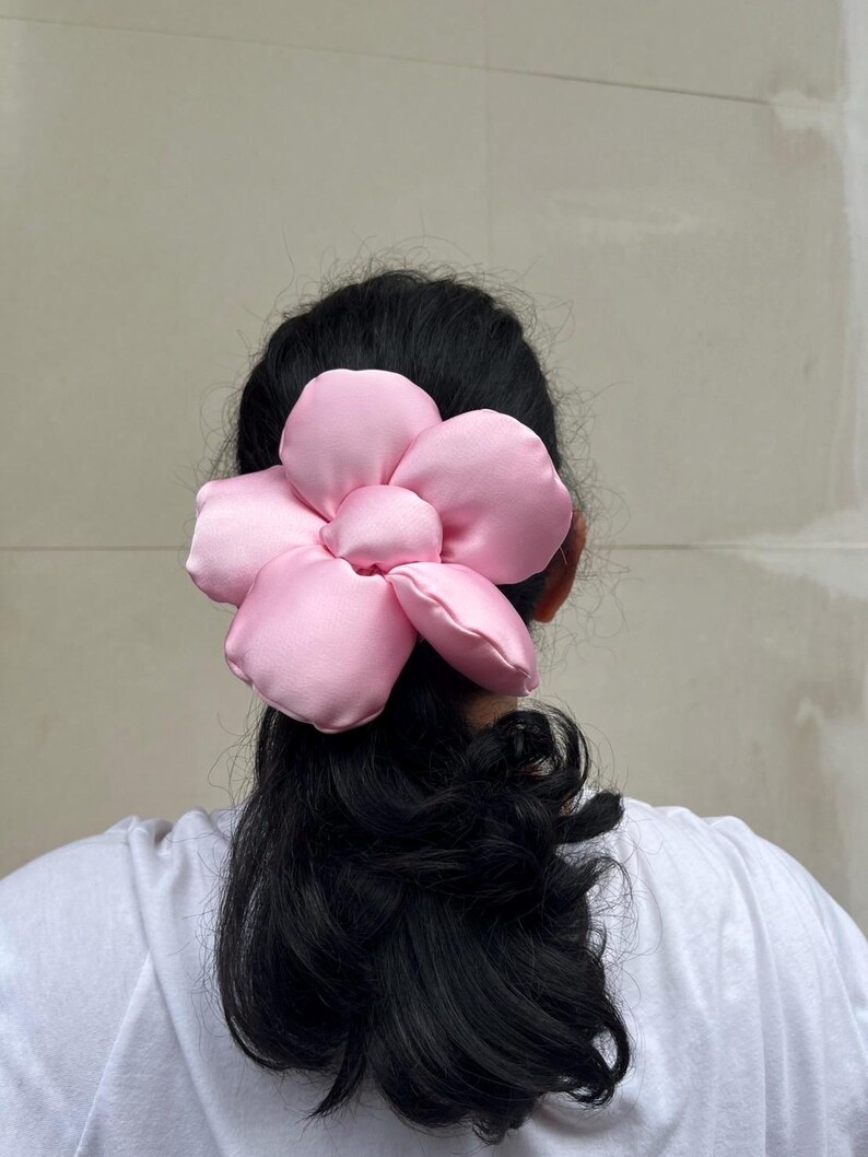 May include: A large, pink flower scrunchie. The hair scrunchie is made of a soft, satin-like material. The flower has multiple petals and a rounded center. The scrunchies is worn on the back of a person's head, with black hair visible. Handmade Fluffy Floral Scrunchie: Long Hair Tie