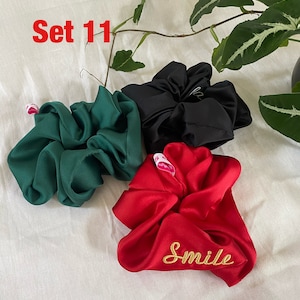 Puede incluir: Conjunto de tres scrunchies en verde, negro y rojo. El scrunchie rojo tiene la palabra "Smile" bordada.