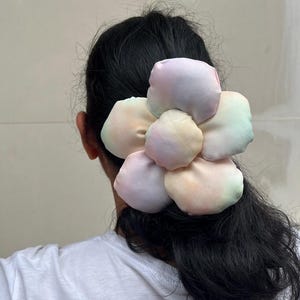 A large flower scrunchie with multiple petals in pastel colors. The petals have a gradient effect, blending shades of pink, yellow, and green. The scrunchies is attached to the back of a person's head, with their dark hair visible. Handmade Fluffy Floral Scrunchie: Long Hair Tie