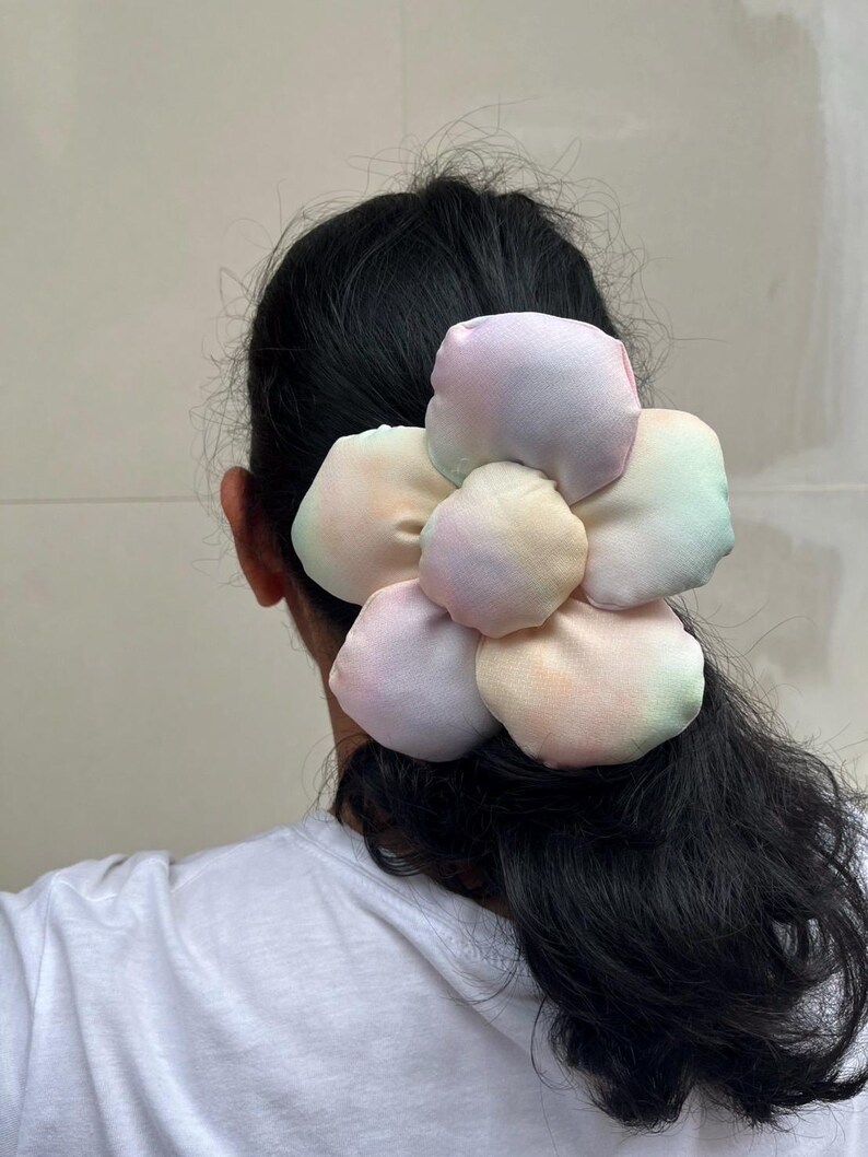A large flower scrunchie with multiple petals in pastel colors. The petals have a gradient effect, blending shades of pink, yellow, and green. The scrunchies is attached to the back of a person's head, with their dark hair visible. Handmade Fluffy Floral Scrunchie: Long Hair Tie