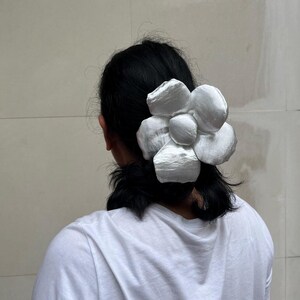 A large, white flower scrunchie. The hair accessory is made of a soft, possibly satin-like material, with multiple layered petals. The flower is attached to a clip, designed to be worn in the hair. The scrunchie is a statement piece. Handmade Fluffy Floral Scrunchie: Long Hair Tie
