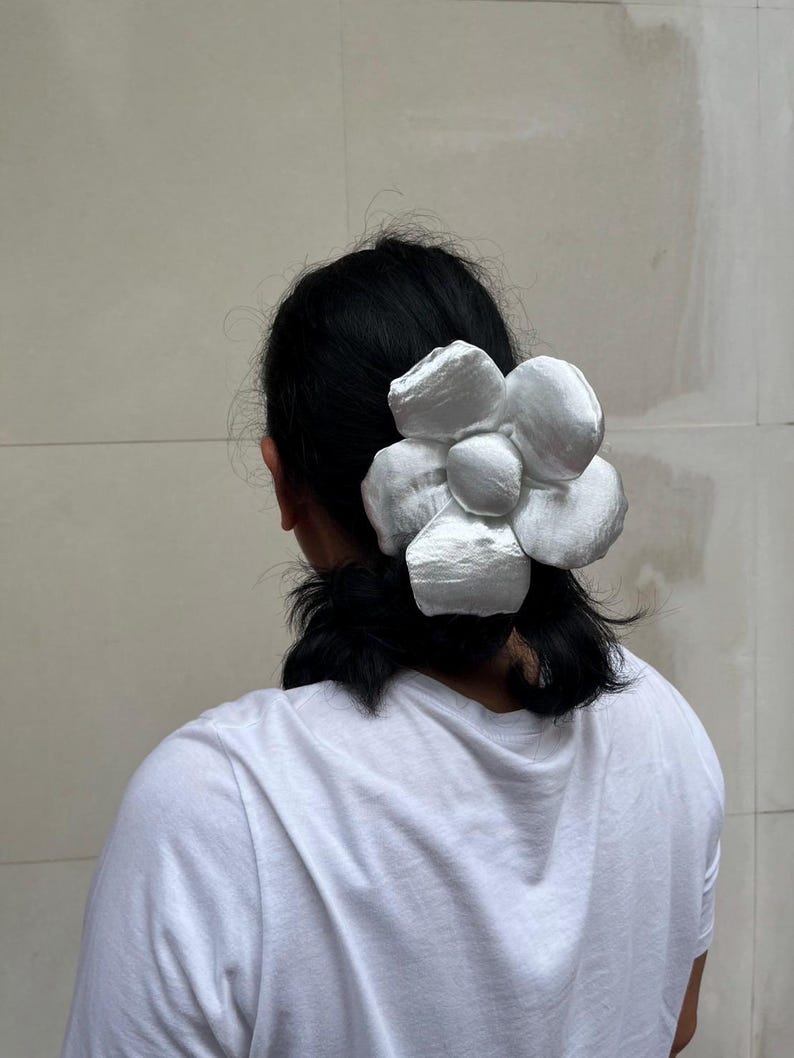 A large, white flower scrunchie. The hair accessory is made of a soft, possibly satin-like material, with multiple layered petals. The flower is attached to a clip, designed to be worn in the hair. The scrunchie is a statement piece. Handmade Fluffy Floral Scrunchie: Long Hair Tie