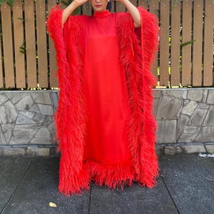 May include: A vibrant red dress with long sleeves and a high neckline. The dress is adorned with fluffy red feathers along the sleeves and hemline. The outfit is a bold fashion statement.