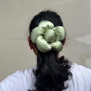 May include: A light green satin flower scrunchie. The hair accessory is in the shape of a large flower with multiple petals. The scrunchie is worn at the back of the head, with dark hair visible. Handmade Fluffy Floral Scrunchie: Long Hair Tie