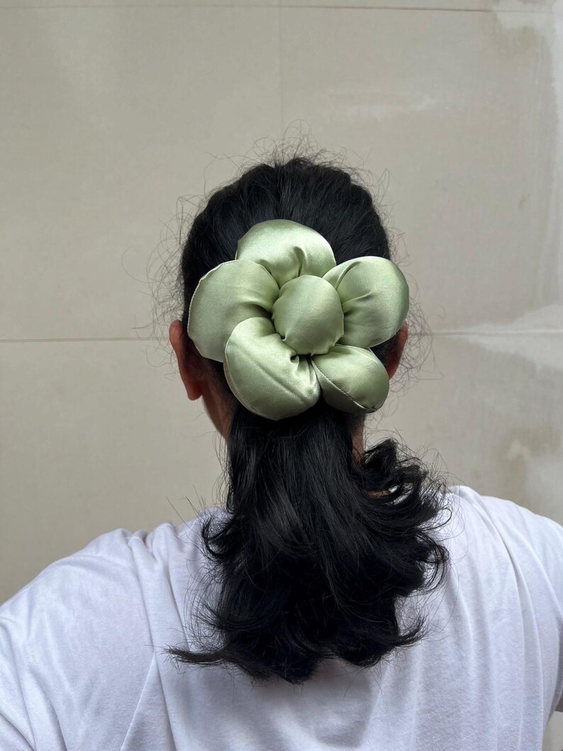 May include: A light green satin flower scrunchie. The hair accessory is in the shape of a large flower with multiple petals. The scrunchie is worn at the back of the head, with dark hair visible. Handmade Fluffy Floral Scrunchie: Long Hair Tie